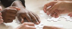 Image of several sets of hands reaching to put together a plain white jigsaw puzzle.